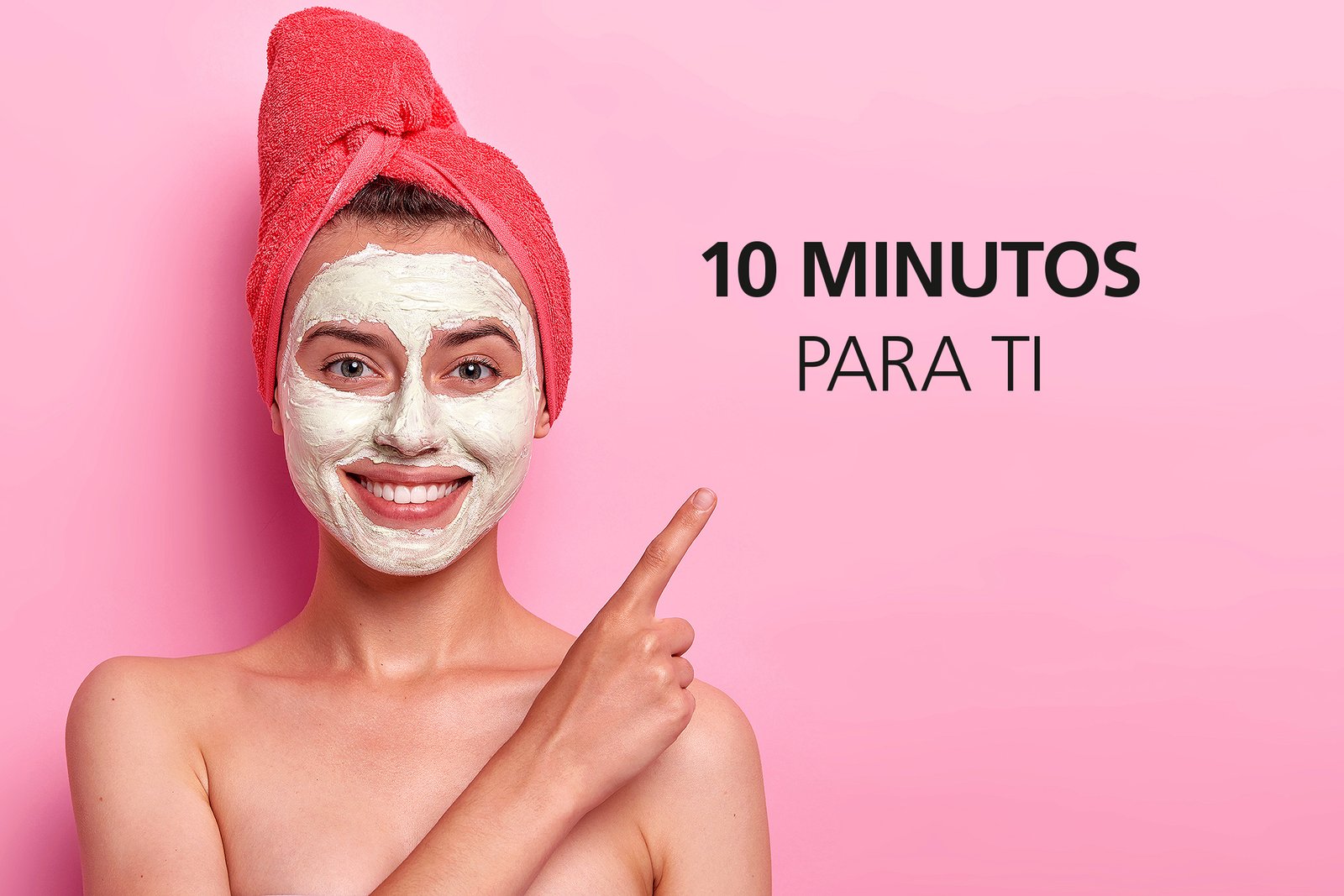 Studio shot of cheerful young woman with gentle smile, points index finger on copy space, advertises beauty item, stands shirtless, wears clay mask and towel poses against pink background. Cosmetology
