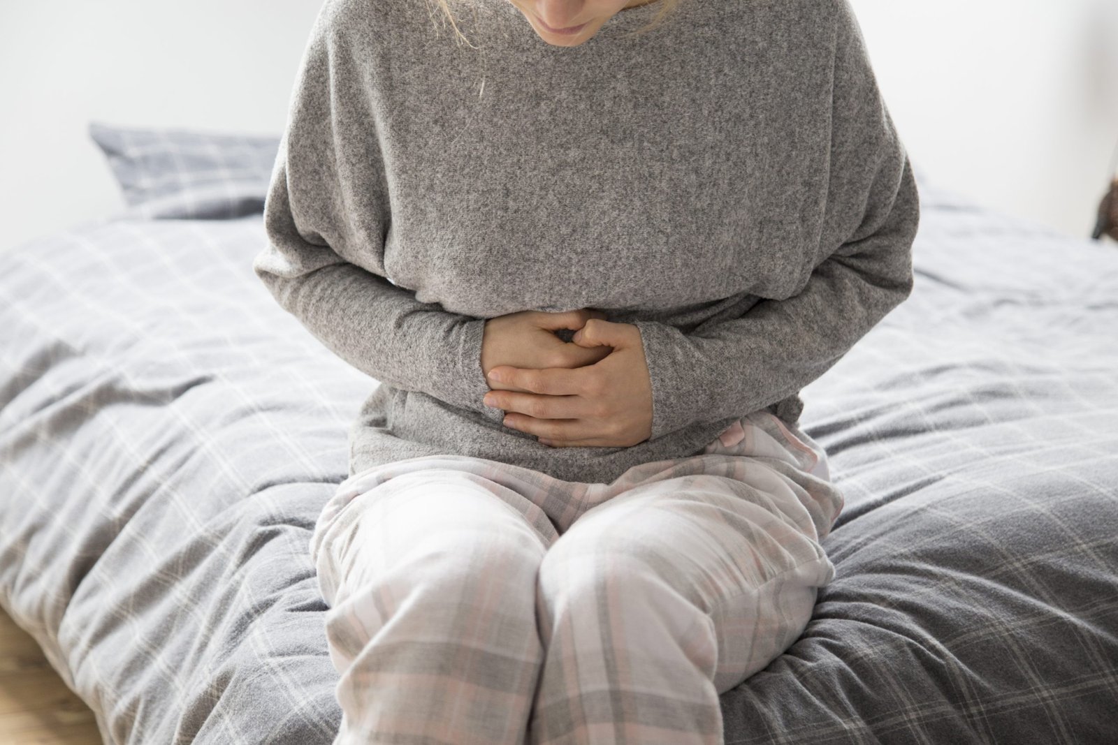 Sick woman in grey homewear sitting on bed, keeping hands on stomach, suffering from intense pain. Illness, stomach ache concept