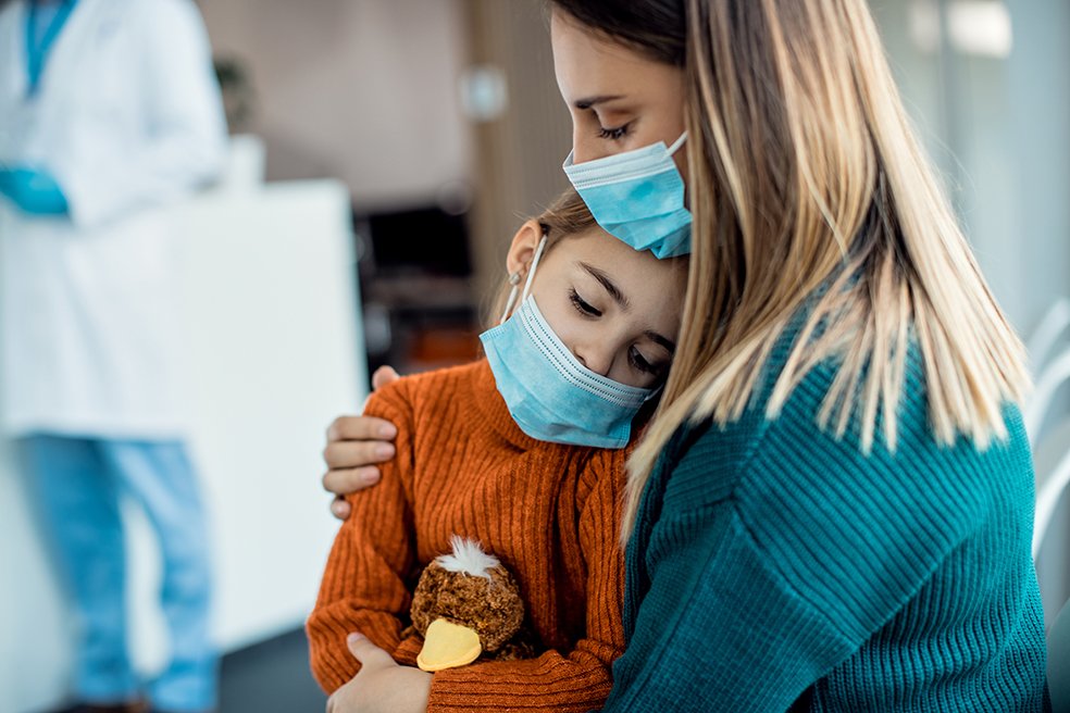 Mother comforting her sad daughter while sitting in waiting room in the hospital and wearing protective face masks.